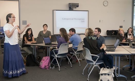 Teacher standing in front of classroom. Students sitting at desks looking at the teacher. 
