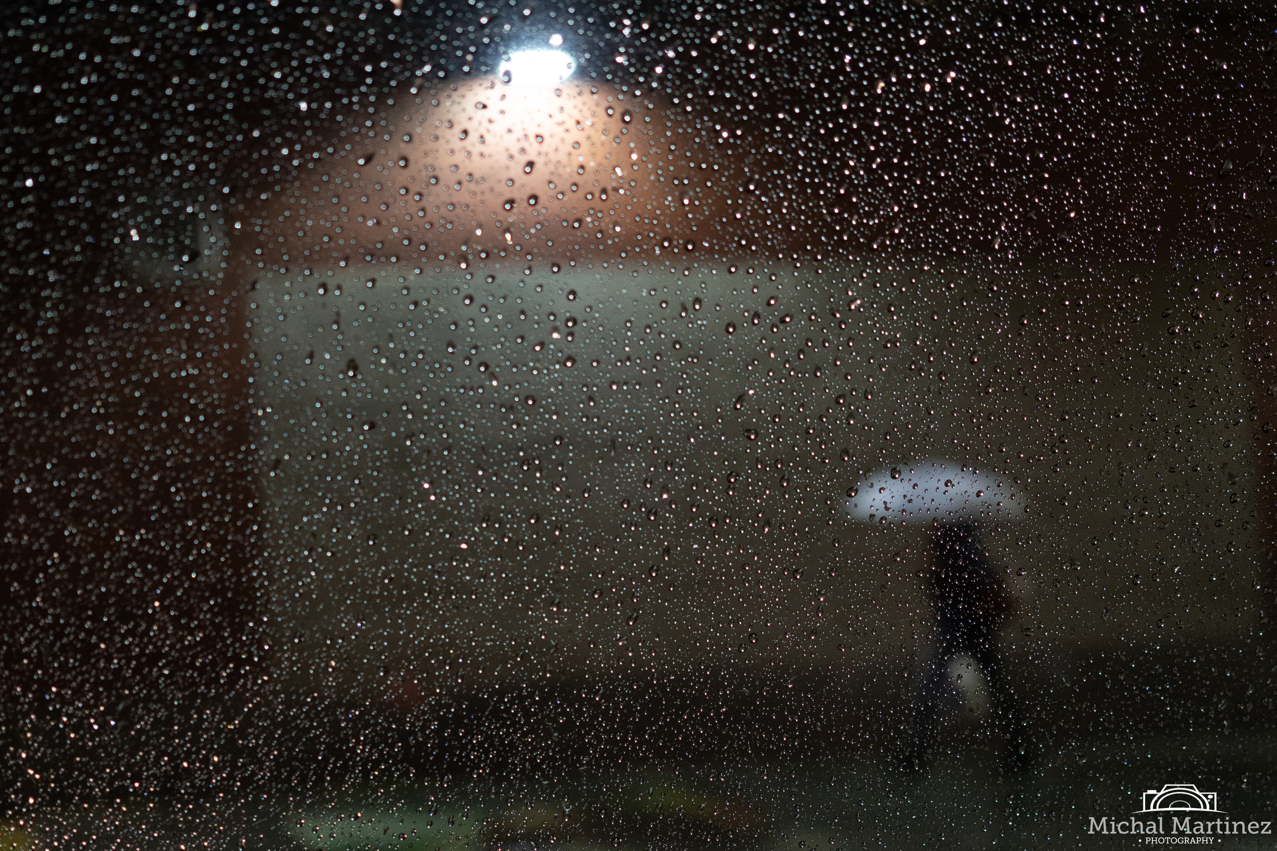 Woman under a white umbrella walking outside by a white garage under a street light. Taken through a car window with rain drops.